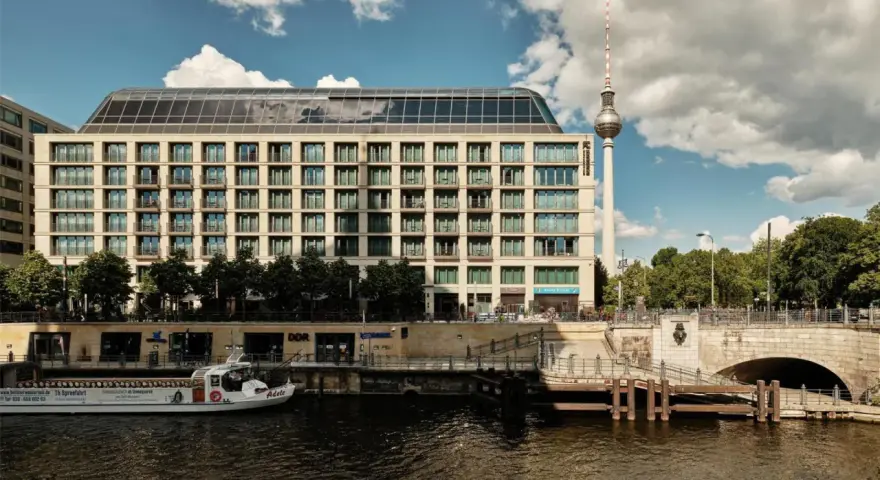 Nowoczesny budynek hotelu Radisson Blu Berlin nad rzeką Sprewą, z widoczną w tle wieżą telewizyjną.