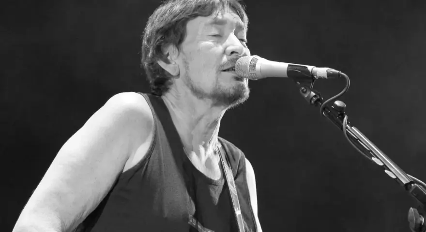 Chris Rea nie żyje. Muzyk śpiewa do mikrofonu na czarnym tle, w czarnej koszulce bez rękawów.