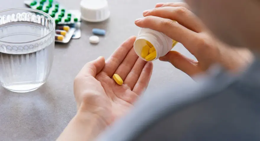 Dłoń wysypująca żółte tabletki z pojemnika na drugą dłoń, obok szklanka wody i opakowania leków.