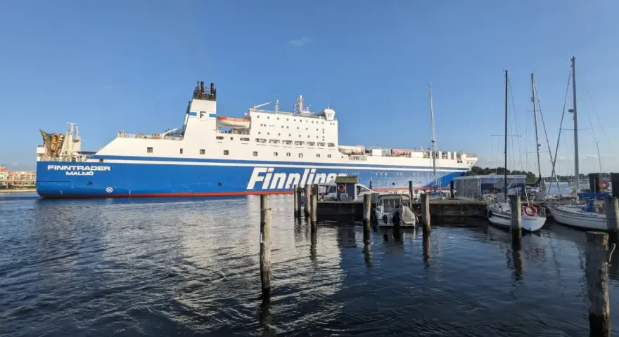 Duży prom Finnlines "Finntrader Malmö" cumuje w porcie, gotowy do rejsu. W tle widać żaglówki. Prom do Finlandii czeka na pasażerów.