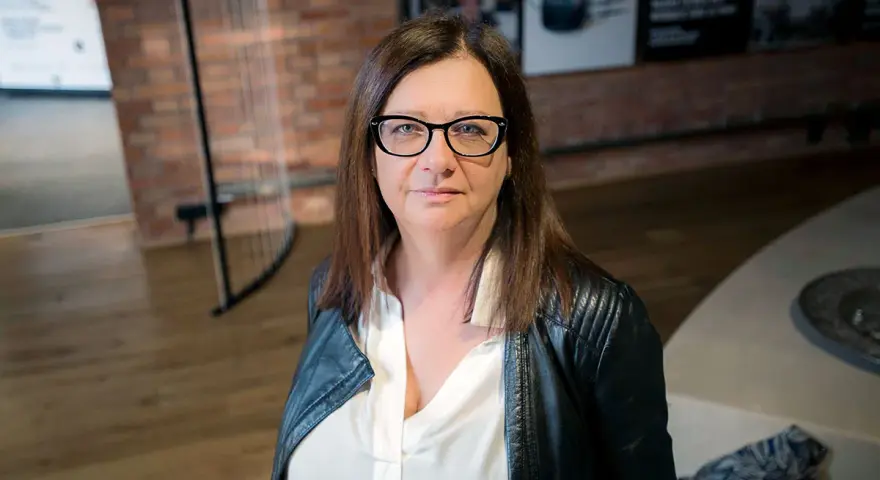 Małgorzata Szczęśniak w okularach i czarnej ramonesce, zamyślona.