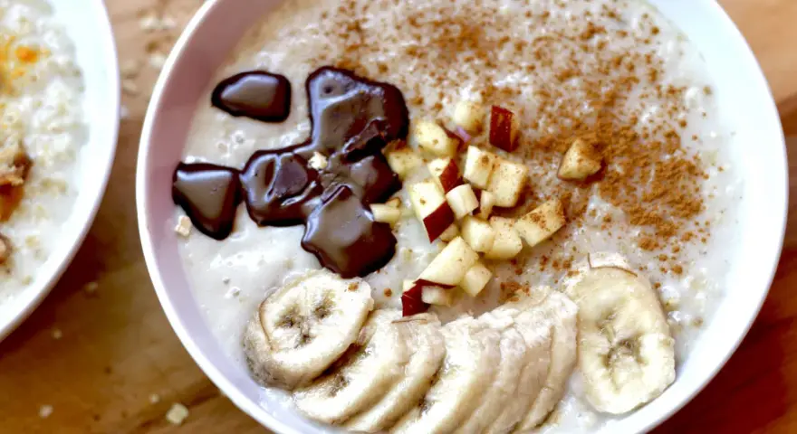 Un tazón de avena cremosa, perfecto para aprender como hacer porridge. Decorado con rodajas de plátano, trocitos de manzana, canela y un toque de chocolate derretido.