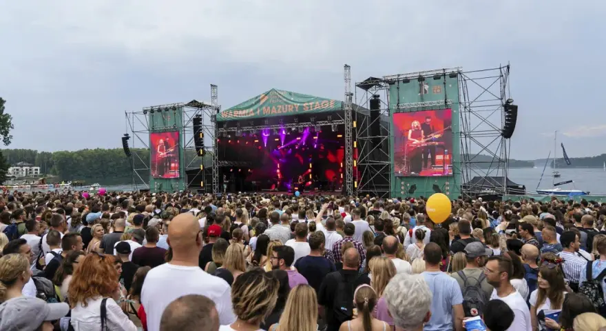 Tłum ludzi na koncercie pod gołym niebem. Na scenie gra zespół, a na ekranach wyświetlany jest green festival line up.
