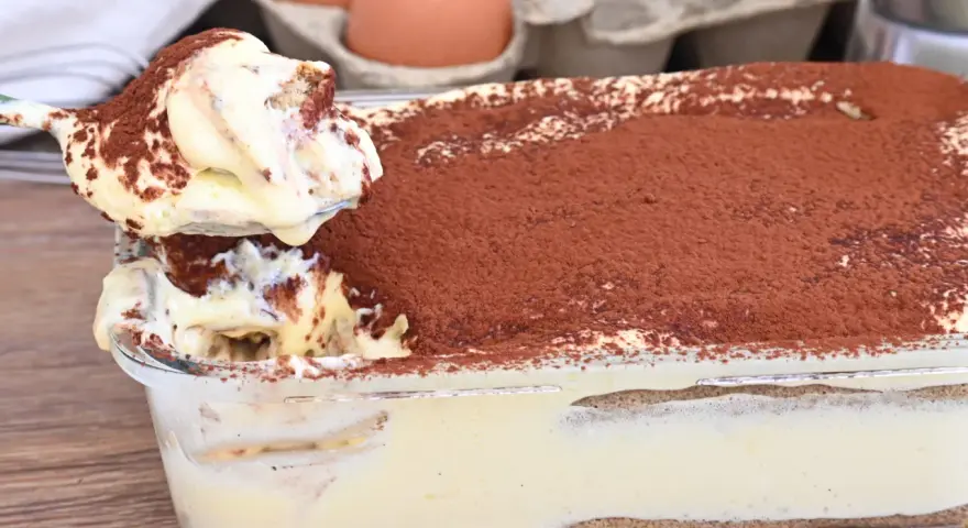 Un cucchiaio preleva una porzione cremosa di tiramisù, con 500 g di mascarpone, cosparso di cacao. Sullo sfondo, uova fresche.
