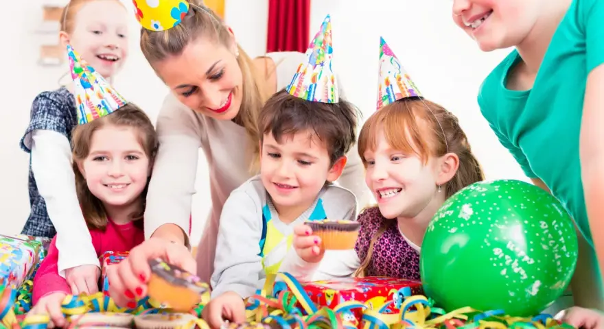Glada barn med kalas-hattar firar ett kalas för en 2-åring med muffins och ballonger.