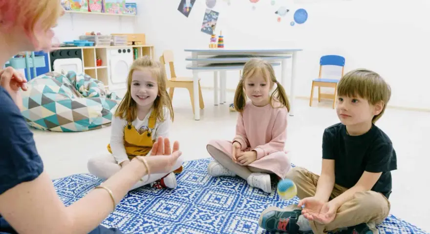 Dzieci siedzą na dywanie, słuchając nauczycielki. To przykład metody dobrego startu, gdzie nauka odbywa się przez zabawę.