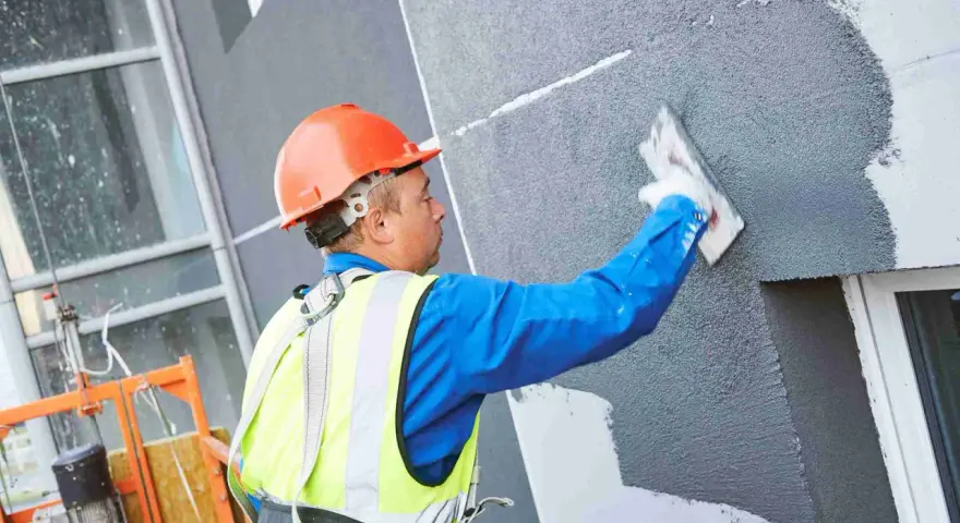 Obrero con casco naranja aplica revestimiento texturizado en fachada, parte de un sistema de aislante térmico exterior.