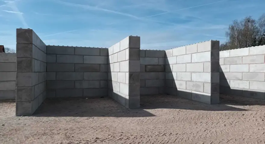 Budowa muru oporowego z bloczków betonowych. Konstrukcja w kształcie litery L, z widocznymi cieniami na piasku.