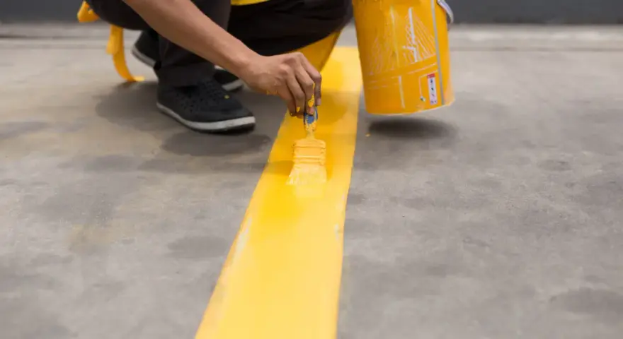 Persona pintando una línea amarilla brillante sobre un suelo de cemento con una brocha.