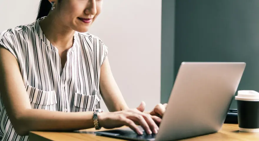 Kobieta uśmiecha się, pracując przy laptopie. Jej przygotowanie pedagogiczne online przebiega sprawnie.