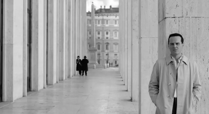 Un uomo in trench osserva l'obiettivo, mentre sullo sfondo due figure in abito scuro passeggiano sotto un colonnato. Potrebbe essere una scena da "Il talento di Mr. Ripley dove è stato girato".