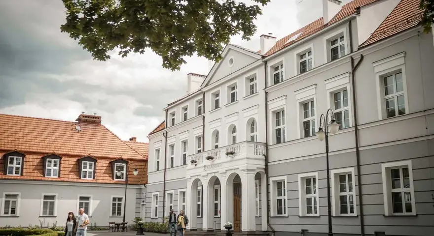 Elegancki pałac Zwartowo z białymi zdobieniami i łukowym wejściem. Na pierwszym planie para spaceruje po brukowanym dziedzińcu.