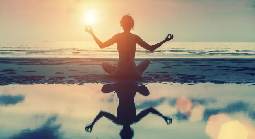 Frau in Lotusposition am Strand bei Sonnenuntergang. Ihre Meditation stärkt das Selbstbewusstsein.