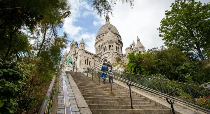 Dwie osoby wspinają się po schodach na wzgórze Montmartre, w tle Bazylika Sacré-Cœur.