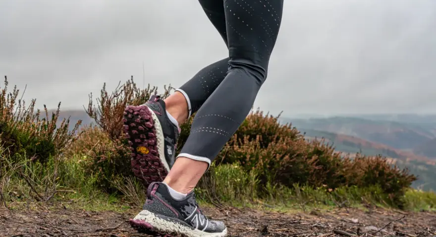 Gambe di una donna che fa trail run su un sentiero fangoso, con erica e colline nebbiose sullo sfondo.