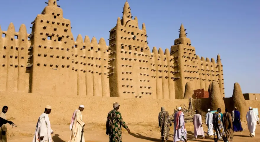 La grande mosquée de Djenné, chef-d'œuvre d'architecture en terre, se dresse sous un ciel bleu. Des hommes en tenues traditionnelles s'approchent, ajoutant vie à ce site historique.