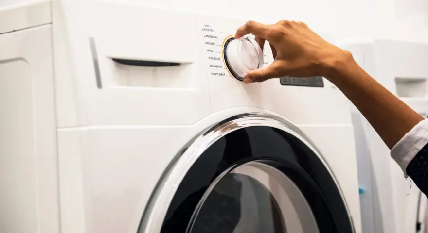 Main d'une personne ajustant le sélecteur d'un sèche-linge, choisissant un programme comme "Coton" ou "Laine". Les dimensions de la machine à laver sont standard.