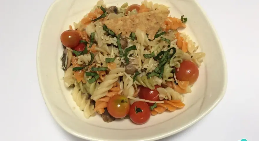 Pasta tricolor con tomates cherry, albahaca y queso rallado en un plato blanco.
