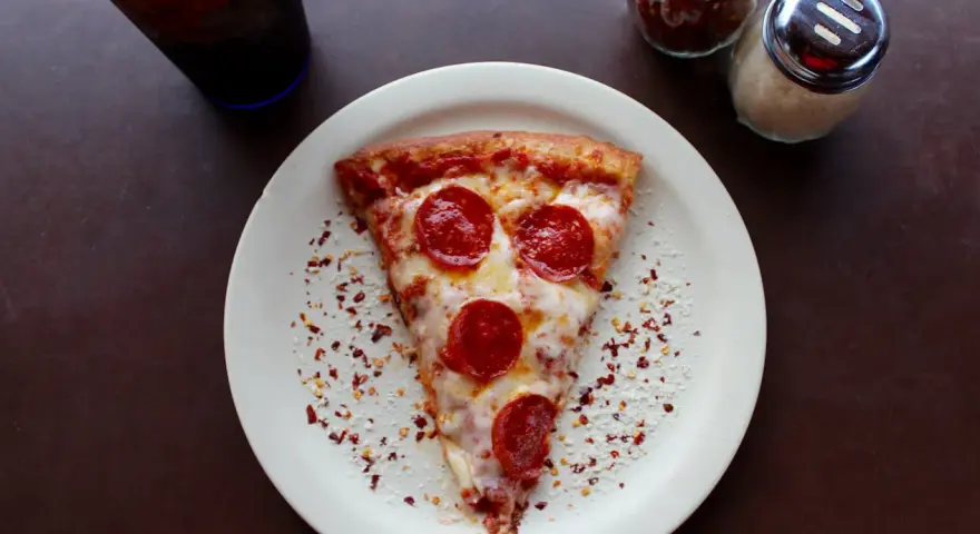 Kawałek pizzy pepperoni na talerzu, posypany płatkami chili i parmezanem. Idealny na szybki lunch, choć warto pamiętać o jego kcal.