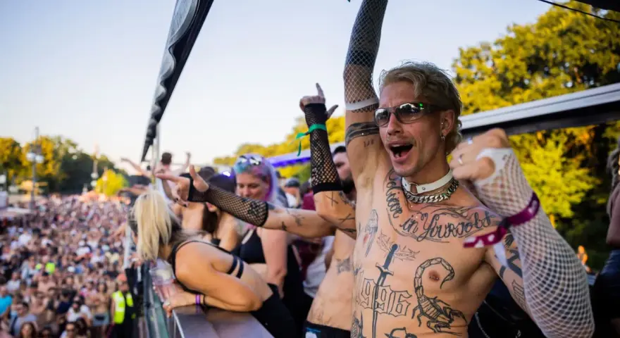 Feiernde Menschenmenge bei einem Festival, die zu **techno music** tanzt. Ein Mann mit Tattoos und Sonnenbrille jubelt.