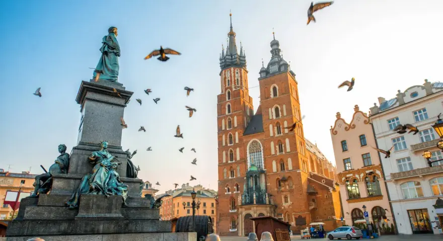 A monument and St. Mary's Basilica dominate this Krakow travel scene. Birds fly around the historic architecture under a clear sky.
