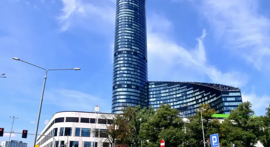 Wysoki, szklany budynek Sky Tower we Wrocławiu, z pewnością ma wiele pięter. Widać też niższy budynek i drzewa.