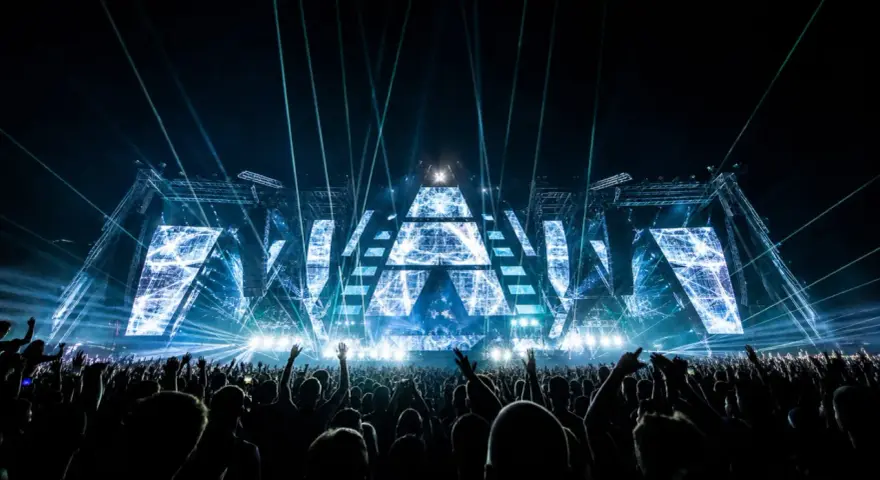 Eine riesige Menge jubelt bei einem EDM-Konzert. Die Bühne ist mit geometrischen Mustern und blauen Lichtern beleuchtet, was die Bedeutung von EDM-Musik für die Fans unterstreicht.