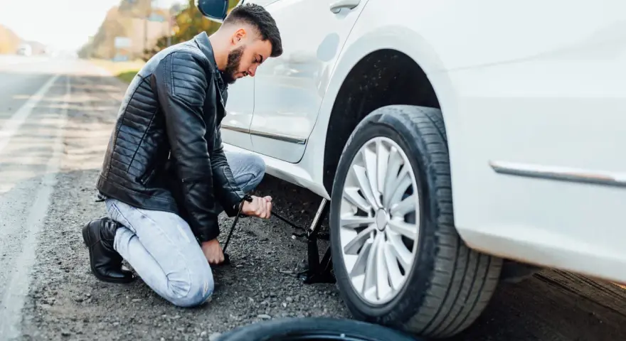 Mężczyzna klęczy przy białym samochodzie, używając podnośnika, by podnieść auto bez kanału. Obok leży zapasowa opona.