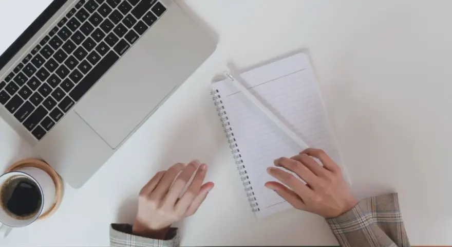 Ręce piszą CV po niemiecku przy laptopie, notatniku i kawie.