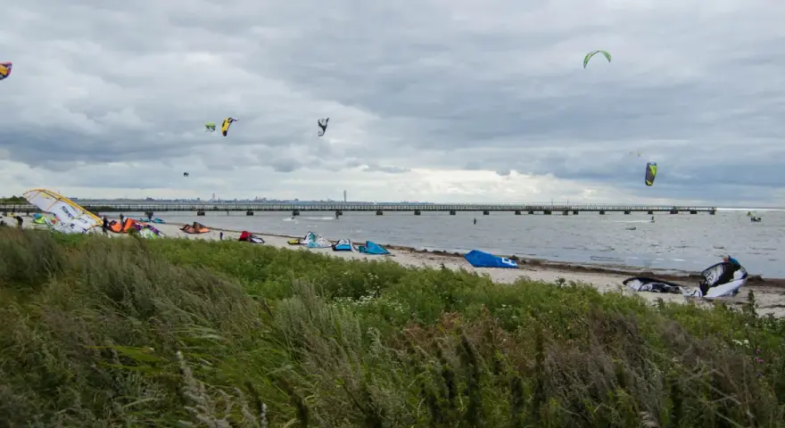 Kitesurfare förbereder sig vid Habo Ljung Camping. Många kites svävar i luften över en strand med en lång pir i bakgrunden.