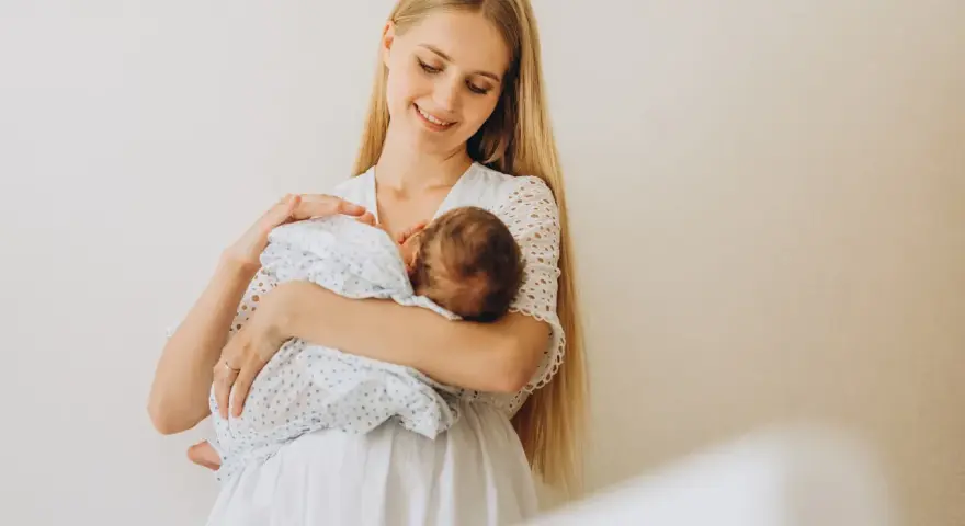 Uśmiechnięta młoda mama tuli noworodka, karmiąc go piersią. Delikatne spojrzenie i biała sukienka podkreślają jej macierzyństwo.