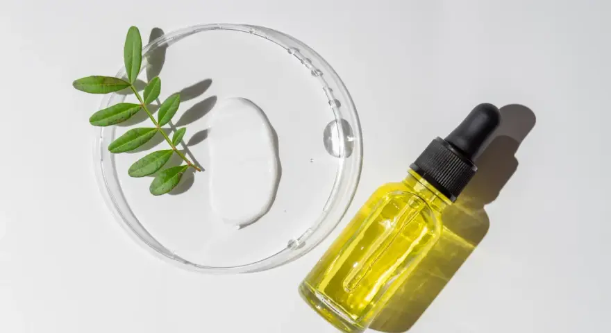 Kropla białego kremu i żółte serum w buteleczce z pipetką, obok gałązka z liśćmi. Składniki do walki z trądzikiem, w tym retinol.