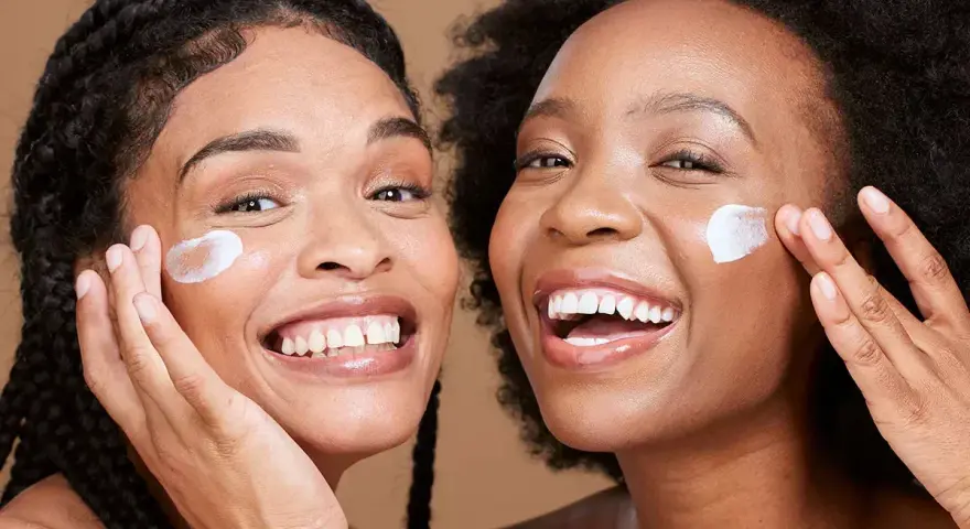 Deux femmes souriantes appliquent une crème sur leur visage, un geste pour une exfoliation visage réussie.