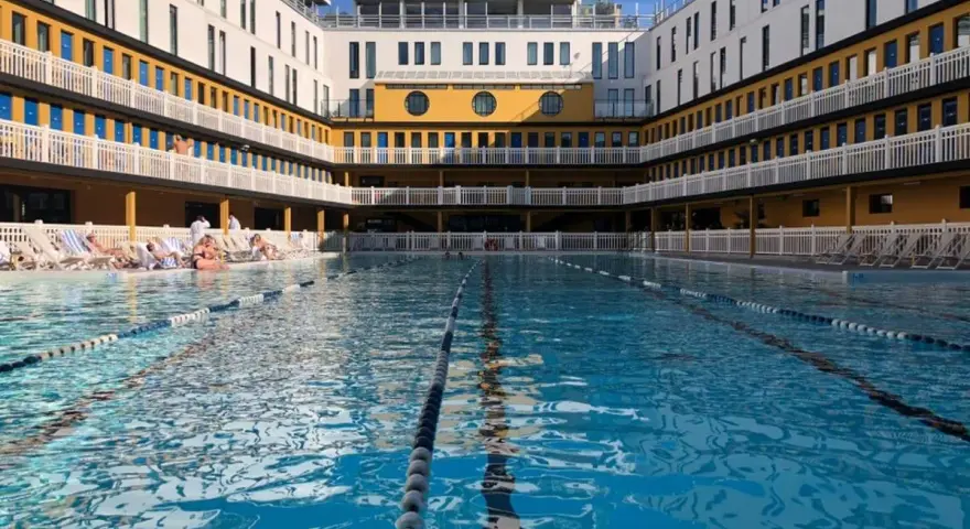 Piscine art déco parisienne, avec ses bâtiments jaunes et blancs, ses balcons et ses transats. L'eau bleue invite à la baignade.