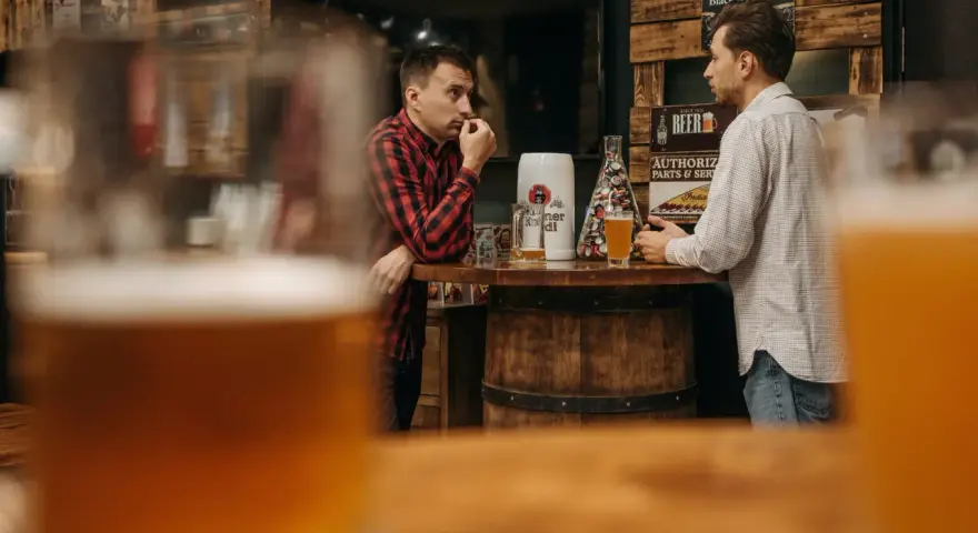 Dwóch mężczyzn rozmawia przy barze z piwem, w tle dekoracje związane z piwem.