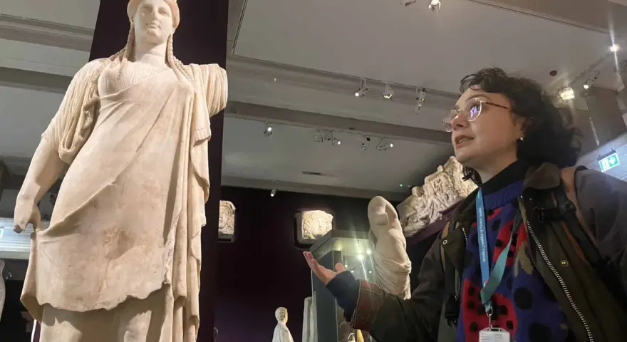 A woman with a museum pass for Turkey gestures towards a marble statue of a woman in a museum.