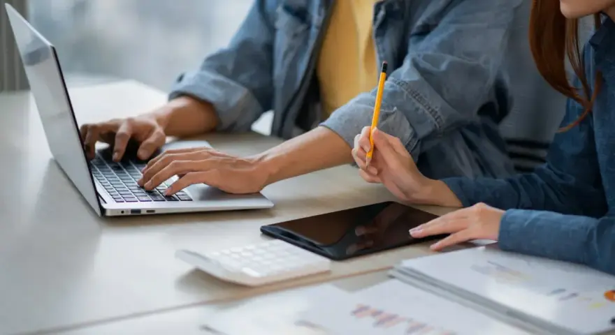 Dwie osoby pracują przy biurku, jedna pisze na laptopie, druga trzyma ołówek. Obok leży tablet i kalkulator. Może to być praca za najniższą krajową za godzinę.