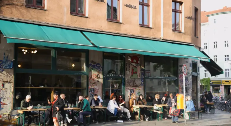 Menschen sitzen an Tischen vor einem Café in Berlin. Die Fassade ist mit Graffiti verziert und trägt die Aufschrift "dorle berlin".