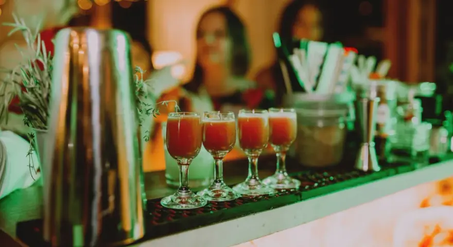 Cztery kieliszki z czerwonym drinkiem stoją na barze, gotowe do podania na weselu. W tle widać gości i dekoracje.