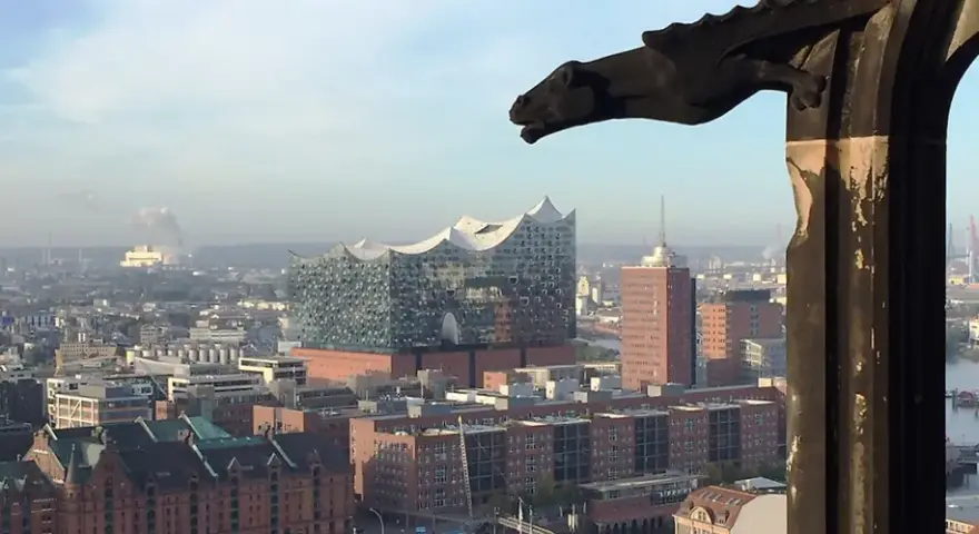 Blick über Hamburg mit der Elbphilharmonie, gesehen durch ein Fenster des Mahnmals St. Nikolai. Ein Wasserspeier wacht über die Stadt.