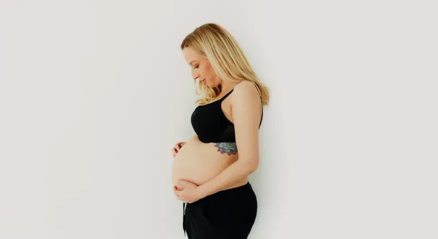 Kobieta w czarnym biustonoszu i spodenkach delikatnie obejmuje swój brzuch w 3 miesiącu ciąży.