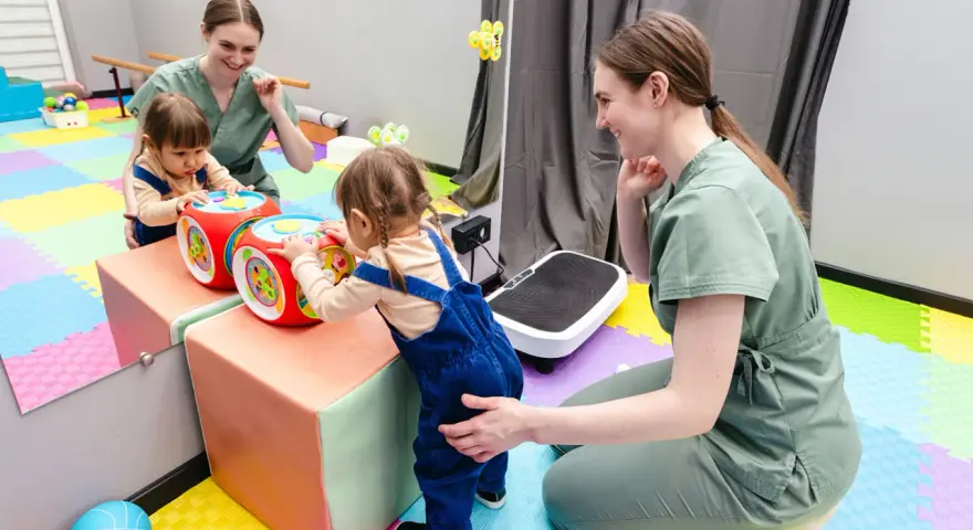 Dwie małe dziewczynki bawią się interaktywną kostką, a fizjoterapeutka wspiera jedną z nich. To może być część terapii wwr co to.