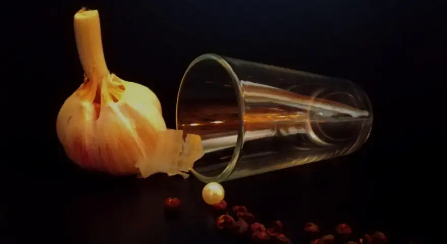 Photographie nature morte : une tête d'ail, un verre renversé, une perle et des baies de poivre rouge sur fond sombre.