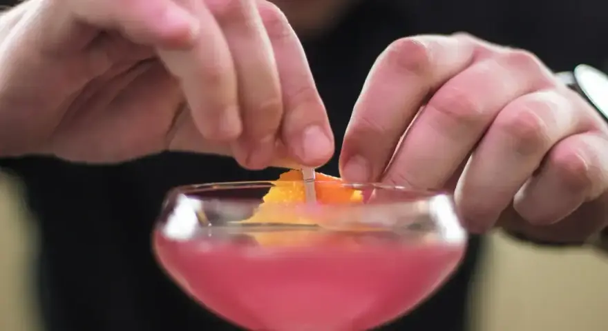 Dłonie barmana ozdabiają drinka skórką pomarańczy. To klasyczny cosmopolitan, gotowy na wieczór w wielkim mieście.