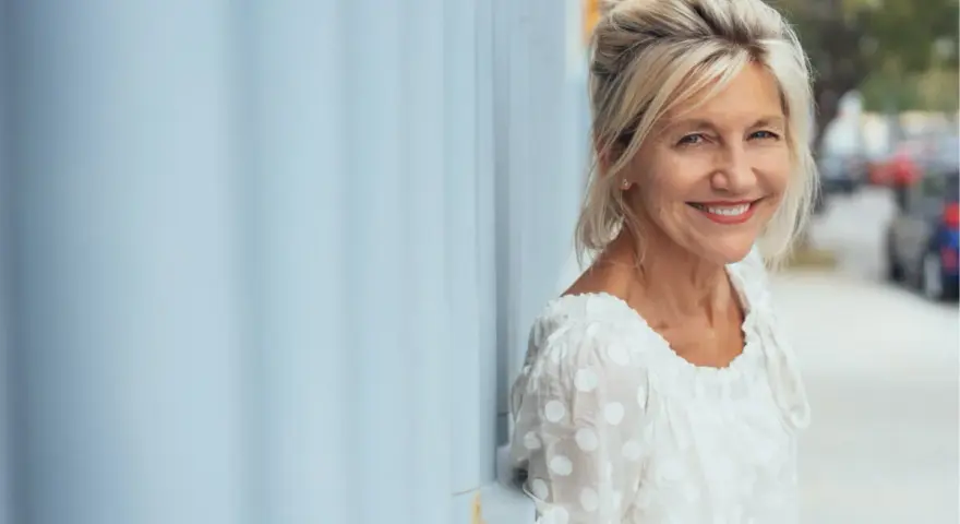 Donna sorridente appoggiata a un muro blu. Potrebbe beneficiare di acido folico in menopausa per il suo benessere.