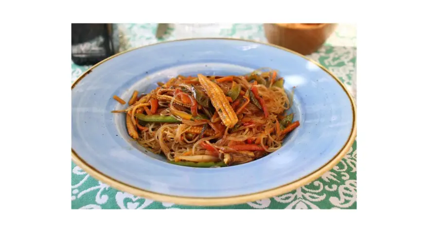 Un piatto di spaghetti di riso con verdure colorate, tra cui peperoni, carote e pannocchiette, condito con una salsa saporita.