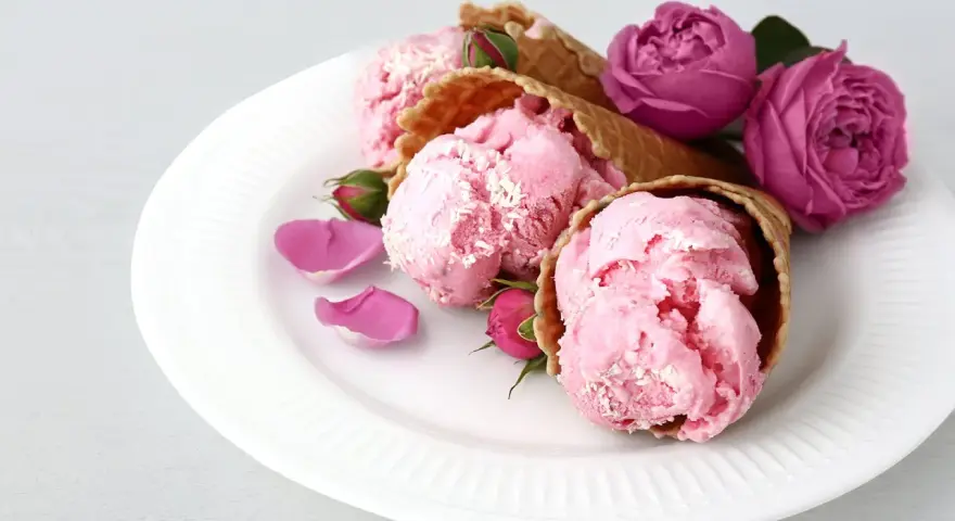 Drei Eiswaffeln mit rosa rosen eis, garniert mit Rosenblättern und Knospen auf einem weißen Teller.