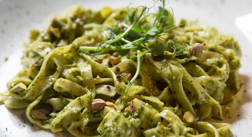 Makaron tagliatelle z zielonym sosem pesto, posypany pistacjami i udekorowany kiełkami.