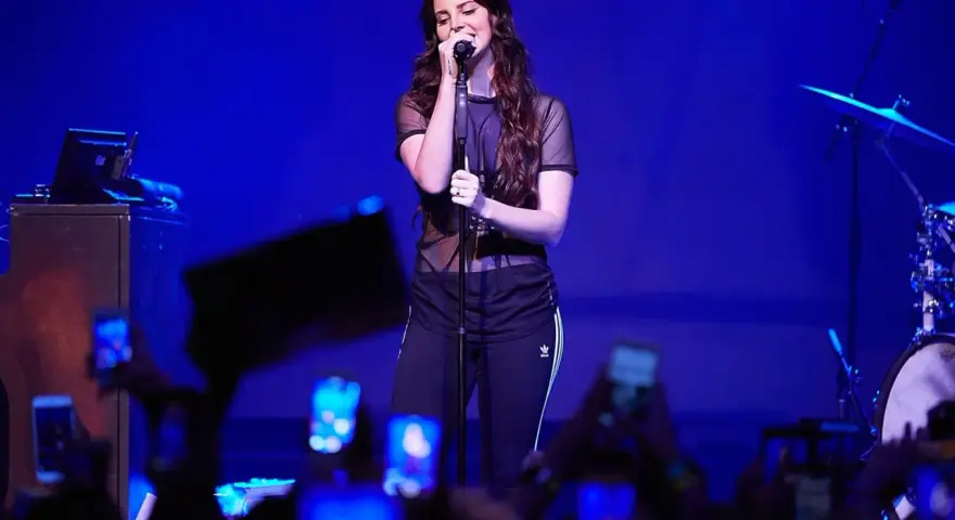 Lana Del Rey, w czarnej, prześwitującej bluzce i sportowych spodniach, śpiewa na scenie. Fani z telefonami tworzą tłum. To jej lana del rey opener.