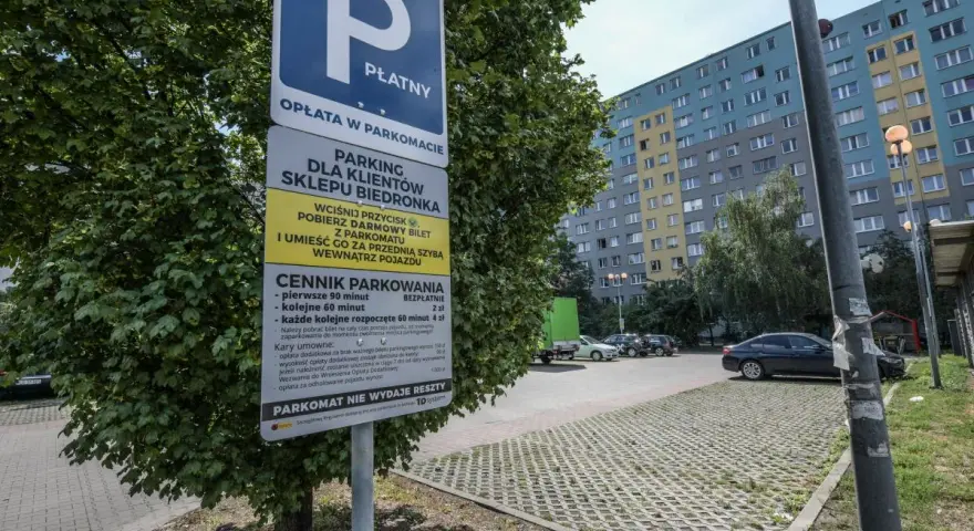 Płatny parking dla klientów Biedronki. Obok drzewa stoi znak informujący o cenniku i zasadach parkowania.